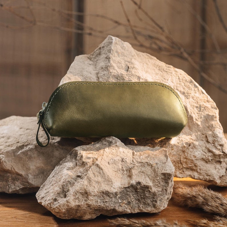 May include: Olive green leather pencil case with a zipper closure and a small loop. The case is resting on a large, textured rock. The stitching is visible along the top seam. The background is a blurred, neutral tone.