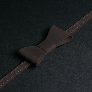May include: A dark brown leather bow tie with a matching strap, stitched with a lighter thread. The bow tie is centered on a dark background, creating a minimalist aesthetic.