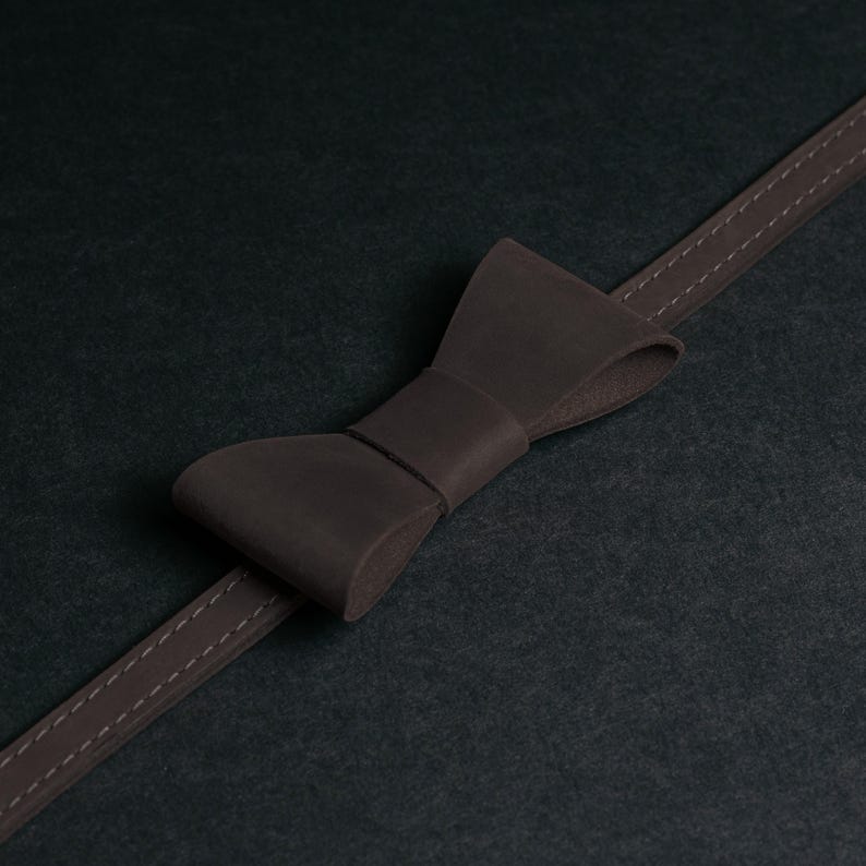 May include: A dark brown leather bow tie with a matching strap, stitched with a lighter thread. The bow tie is centered on a dark background, creating a minimalist aesthetic.