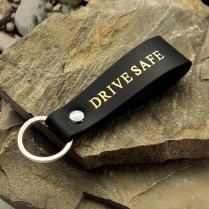 Può includere: Portachiavi in pelle nera con la scritta "DRIVE SAFE" in lettere dorate. Il portachiavi ha un anello in metallo argentato e un rivetto argentato. Il portachiavi è appoggiato su una superficie rocciosa ruvida color beige.