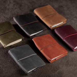 Puede incluir: Una colección de cuadernos de cuero en varios colores, incluyendo verde oliva, negro, marrón claro, burdeos, gris y óxido. Cada cuaderno está asegurado con una banda elástica. Los cuadernos están dispuestos sobre una superficie oscura y texturizada.