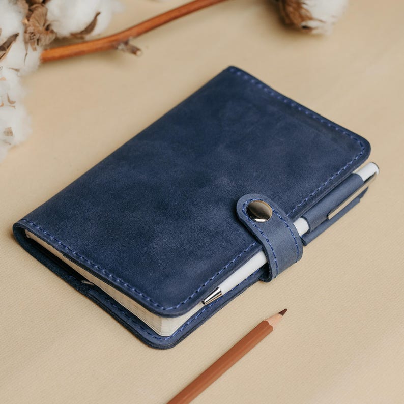 May include: A blue leather notebook with a pen holder and a snap closure. The notebook is lying on a beige surface.