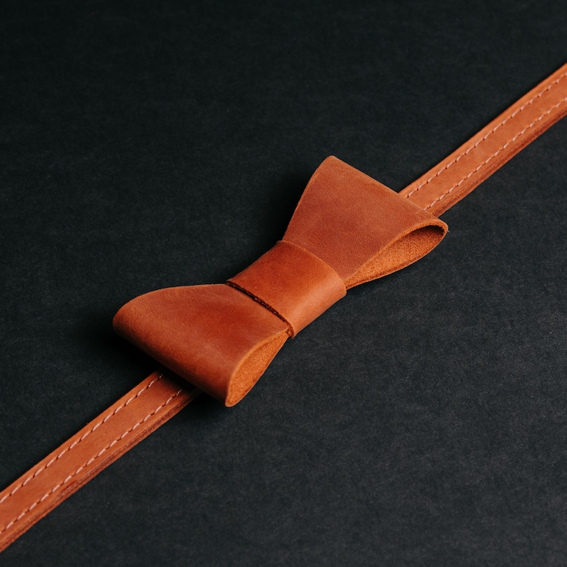 May include: A brown leather bow tie and strap, displayed on a dark backdrop. The bow tie is made from folded leather, with a band around the middle. The strap is stitched with a lighter thread, showcasing the craftsmanship.
