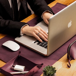 May include: A person using a silver laptop on a purple desk mat. A matching leather tray holds a white charger and pen. A white mouse and a small succulent in a gray pot are also on the desk. A small purple zipped pouch is on the right.