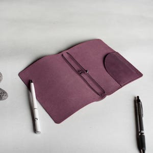 May include: A plum-colored leather journal cover with a pocket and elastic closure. A white pen and a black pen are placed on the surface next to the open journal. The journal cover is open, revealing the interior.