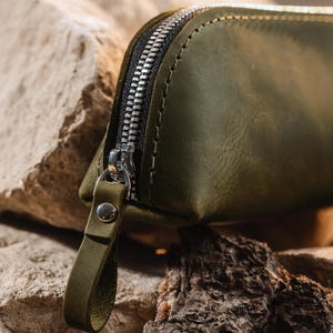 May include: Close-up of an olive green leather pouch with a silver zipper. The pouch has a small loop attached to the zipper pull. The background features rough, textured rocks and wood.