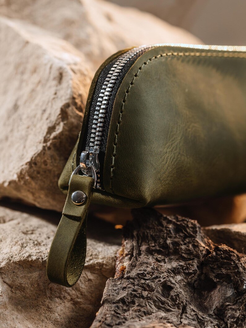May include: Close-up of an olive green leather pouch with a silver zipper. The pouch has a small loop attached to the zipper pull. The background features rough, textured rocks and wood.