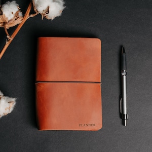 May include: A brown leather planner with a black elastic band and the word "PLANNER" embossed on the front. A silver pen is lying next to the planner on a black surface.