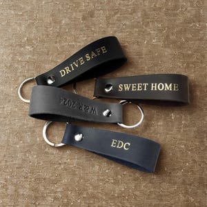 May include: Four leather keychains with gold lettering. The keychains are black, brown, and gray. The text on the keychains reads "Drive Safe", "Sweet Home", "W&R 2024", and "EDC".