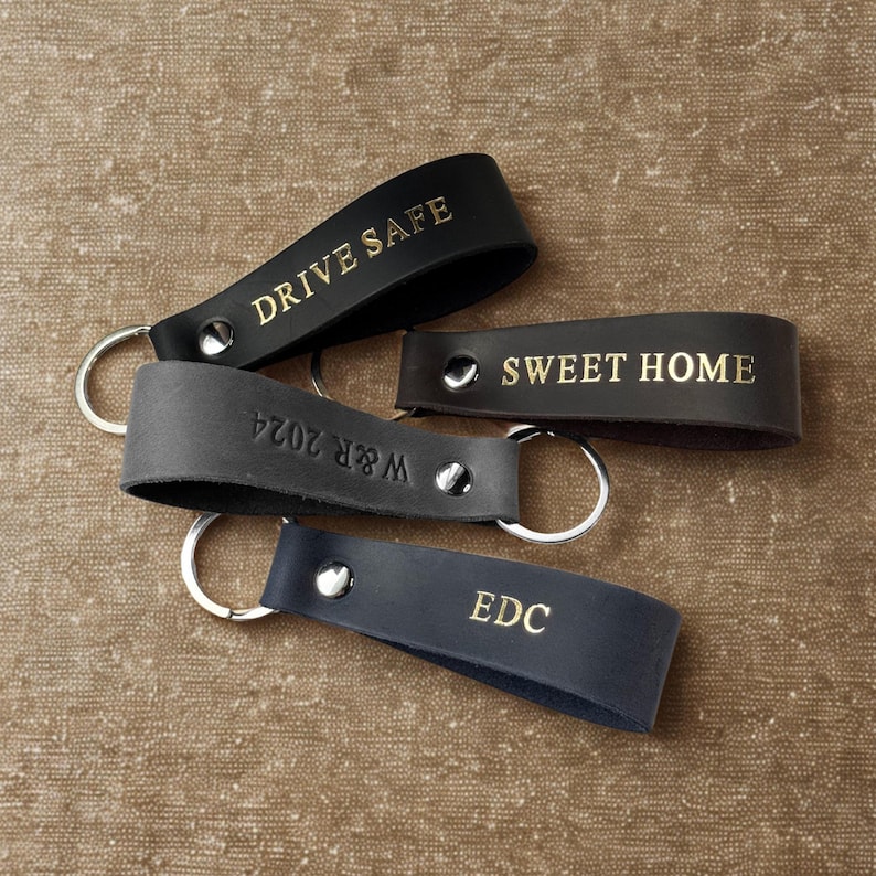 May include: Four leather keychains with gold lettering. The keychains are black, brown, and gray. The text on the keychains reads "Drive Safe", "Sweet Home", "W&R 2024", and "EDC".