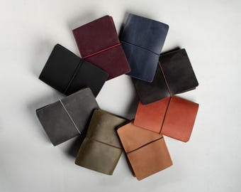 May include: A circular arrangement of eight leather journals in various colours including black, dark grey, olive green, tan, orange, dark brown, navy blue, and burgundy, each secured with a thin elastic band wrapped vertically around the cover.