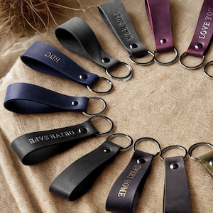 May include: A collection of leather keychains in various colors including black, navy, gray, and purple, arranged in a circular pattern. Each keychain features a metal rivet and a silver-toned split ring, with some engraved with text like W&R 2024, EDC, DRIVE SAFE, LOVE YOU, and SWEET HOME.