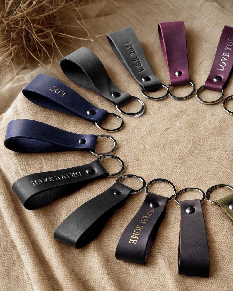 May include: A collection of leather keychains with different engraved messages, including "Love You", "Drive Safe", "Sweet Home", "We're 2024", and "BDC". The keychains are arranged in a circle on a brown burlap background.