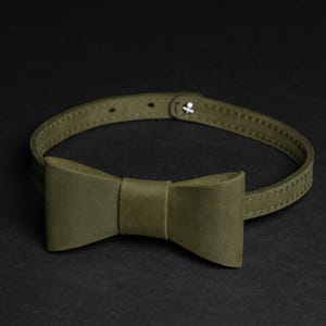 May include: An olive green leather bow tie collar with an adjustable strap. The bow tie is centrally positioned on the collar and has a traditional bow shape. The collar fastens with a silver-coloured snap closure. The background is black.