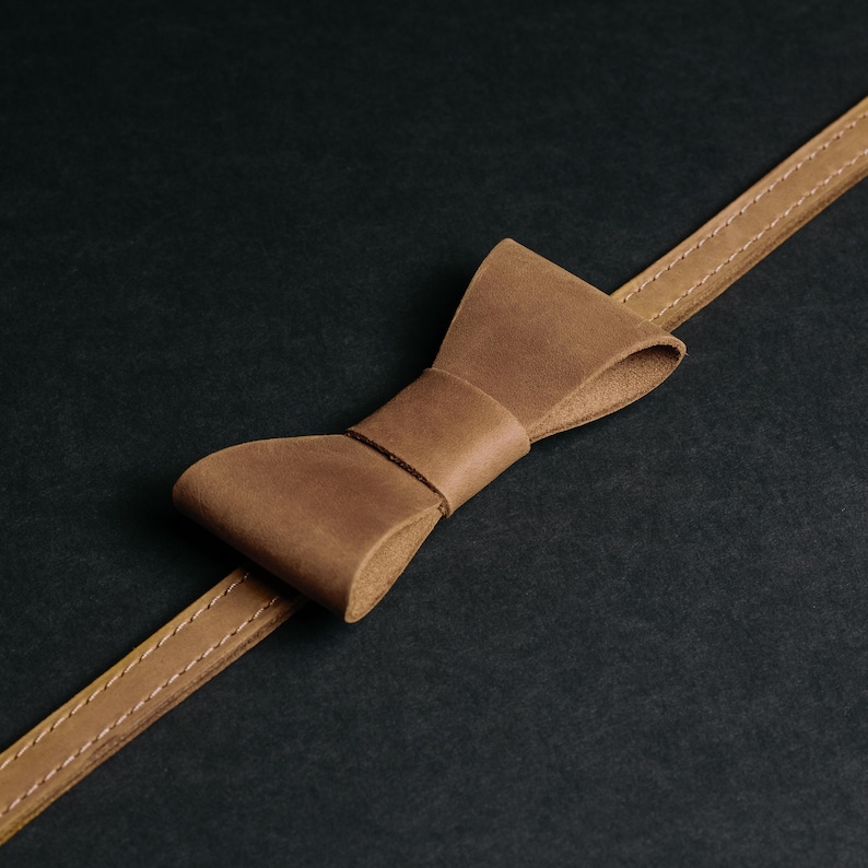 May include: A brown leather bow tie with a stitched strap. The bow tie is made of folded leather and is secured with a thin, dark brown band. The strap is a lighter shade of brown and has visible stitching.