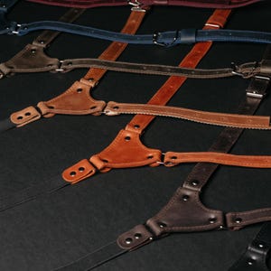 May include: A collection of leather suspenders in various colors including tan, brown, dark brown, blue, and burgundy. Each pair features a triangular leather back piece, metal rivets, adjustable straps with metal buckles, and elastic ends.