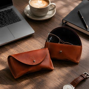 May include: Two tan leather glasses cases with a curved flap and a metal snap closure. One case is closed and features the embossed initials N.E. on the front, while the other is open and contains a pair of dark-rimmed sunglasses inside.