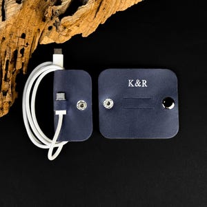 Puede incluir: Dos organizadores de cables de cuero azul marino con cierres de botón a presión plateados. Un organizador contiene un cable blanco. El otro organizador tiene las iniciales "K&R" en letras blancas. El fondo es negro.