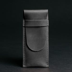 May include: A gray leather case with a flap closure and a strap. The case is rectangular with rounded edges and visible stitching. The background is a dark gray.