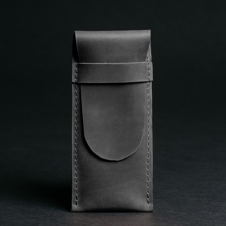 May include: A gray leather case with a flap closure and a strap. The case is rectangular with rounded edges and visible stitching. The background is a dark gray.