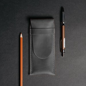 May include: A dark gray leather pen case with a pencil and pen on a black background. The pencil is orange, and the pen is bronze and black. The pen case has a flap closure and stitched edges.