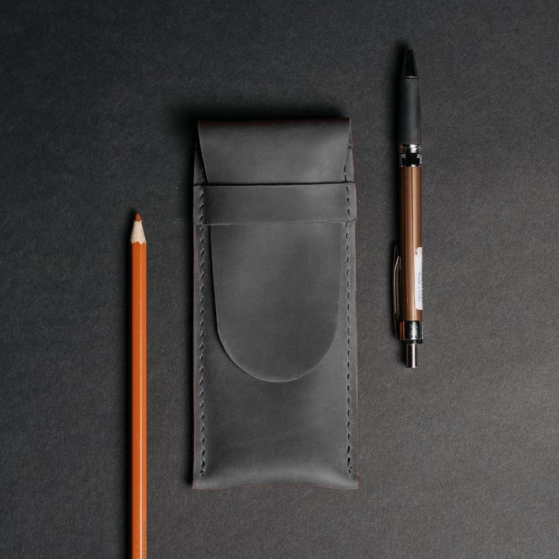 May include: A dark gray leather pen case with a pencil and pen on a black background. The pencil is orange, and the pen is bronze and black. The pen case has a flap closure and stitched edges.