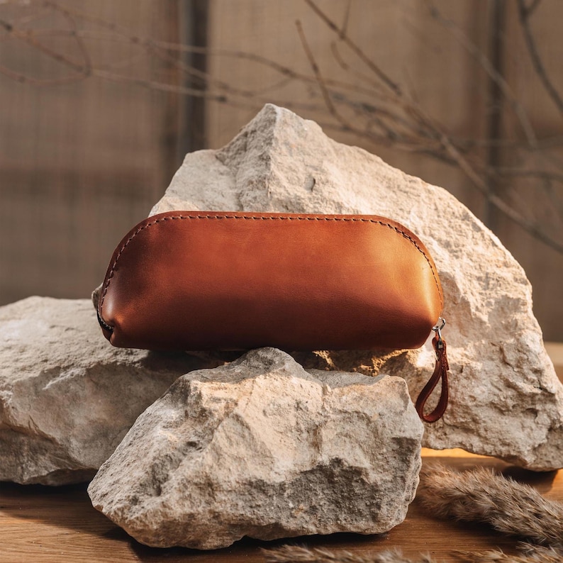 May include: A brown leather pencil case with a zipper closure and a wrist strap. The case is oval-shaped and sits on a rough, light-colored stone surface. The leather has a smooth texture and a warm, reddish-brown color.