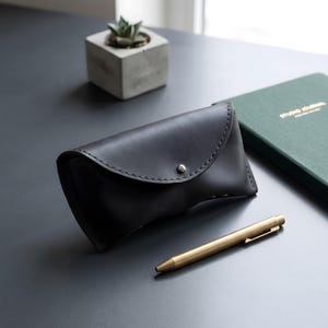 May include: A dark charcoal gray leather glasses case with a curved flap, visible stitching along the edges, and a single silver-toned metal snap button closure. The case is positioned next to a metallic gold-colored pen on a flat surface.