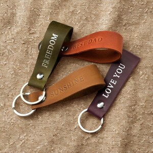 May include: Four leather keychains with metal rings. The keychains are brown, green, purple, and orange. The keychains are engraved with the words "Freedom", "Best Dad", "Sunshine", and "Love You".
