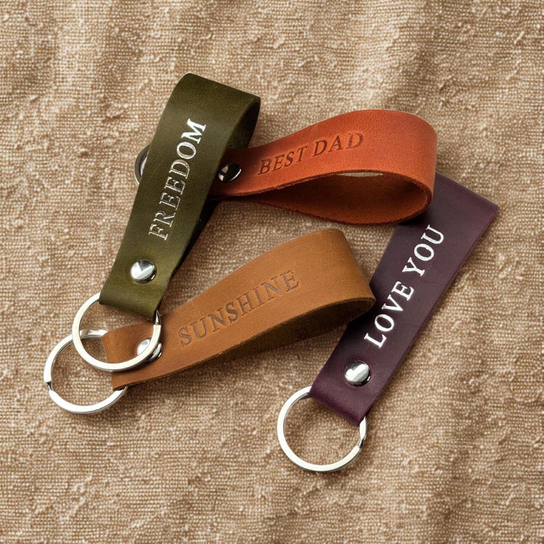 May include: Four leather keychains with metal rings. The keychains are brown, green, purple, and orange. The keychains are engraved with the words "Freedom", "Best Dad", "Sunshine", and "Love You".