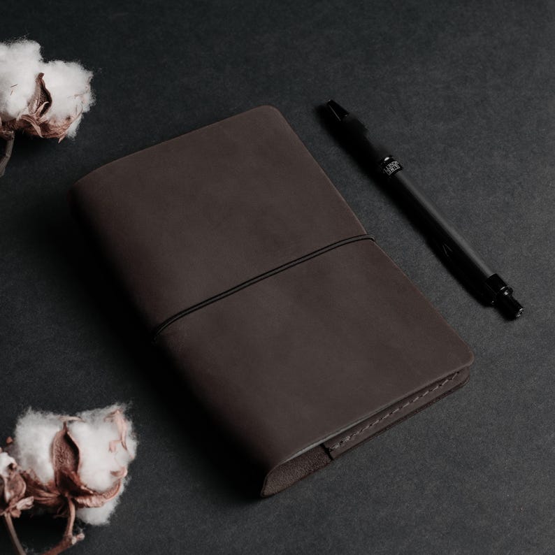 May include: A brown leather bound journal with a black pen on a dark gray surface.