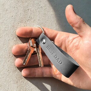 May include: A gray leather keychain with the embossed text "BEST DAD" is attached to a silver key ring with two keys. The keychain is held in a hand against a neutral background.