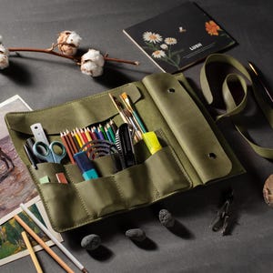 Peut inclure: Un étui à crayons en cuir vert olive ouvert, rempli de crayons de couleur, de pinceaux et d'autres outils. Une aquarelle d'un cheval et un carnet avec des illustrations florales sont également visibles.