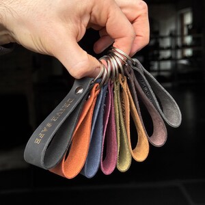 May include: A set of eight leather keychains with different colors and engraved text. The keychains are attached to a silver key ring. The text on the keychains includes "DRIVE SAFE", "SUN", and "ROCK".