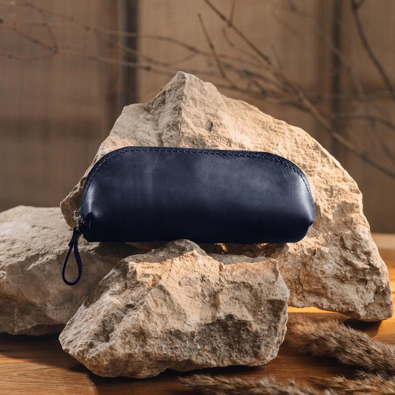 May include: A navy blue leather pencil case with a zipper closure. The case is oval-shaped and rests on a light-colored rock formation. The background features a neutral-toned backdrop and dried foliage.