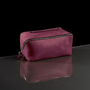 May include: A rectangular, burgundy-colored leather travel bag with a silver zipper closure. The bag has a small handle on top and is reflected in the glossy surface below. The bag is designed for toiletries or other small items.