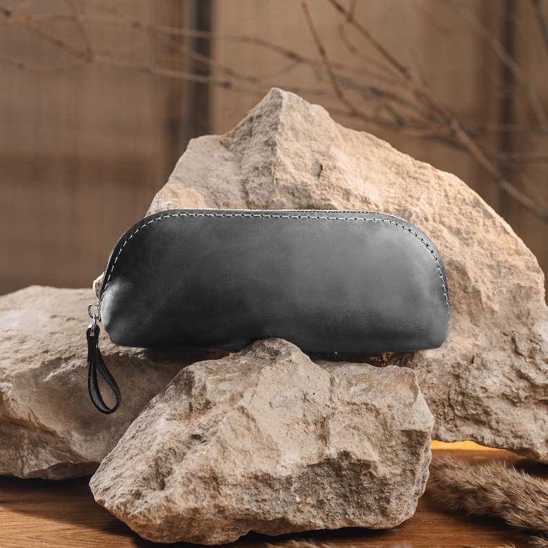 May include: A dark gray leather pencil case with a white stitched zipper closure and a small wrist strap. The case is oval-shaped and rests on a large, rough rock formation. The background is a blurred wooden surface.