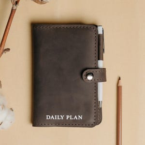 May include: A brown leather daily planner with a pen holder and a silver button closure. The planner has the words "DAILY PLAN" embossed in white on the front.