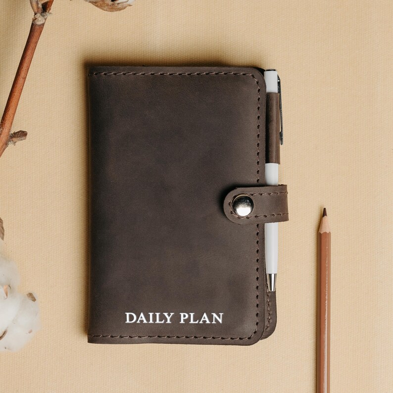 May include: A brown leather daily planner with a pen holder and a silver button closure. The planner has the words "DAILY PLAN" embossed in white on the front.