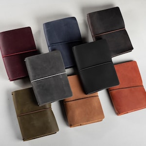 Può includere: Una collezione di diari in pelle in vari colori, tra cui bordeaux, blu navy, nero e marrone. Ogni diario è chiuso con un elastico. I diari sono disposti su una superficie bianca.