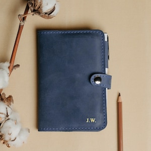 May include: A navy blue leather notebook with a pen holder and a snap closure. The notebook has the initials "J.W." embossed in gold on the front cover.