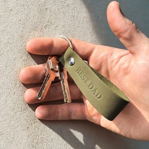 Może przedstawiać: Zielony skórzany brelok do kluczy z srebrnym kółkiem i trzema kluczami. Brelok ma wygrawerowane słowa "BEST DAD".