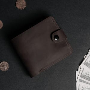 May include: A dark brown leather bifold wallet featuring a snap button closure tab and visible stitching along the edges. The wallet is positioned on a dark surface alongside scattered coins and a stack of United States paper currency bills.