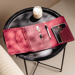 May include: A deep magenta leather journal cover featuring an interior with multiple card slots, a pen loop holding a silver pen, and a vertical pocket containing a postcard with the text LISBOA. The cover includes thin elastic cords for binding.