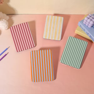 May include: A collection of five rectangular coasters with vertical striped patterns in various colors: red and white, yellow and white, blue and orange, green and white, and a pink and white. The coasters are arranged on a pink surface.