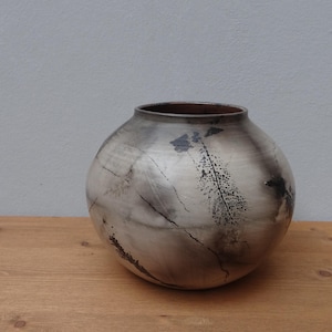 May include: A round, white ceramic vase with a brown rim and black feather-like markings. The vase has a textured surface and is sitting on a wooden surface.