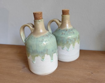 May include: Two ceramic jugs with handles and cork stoppers. The jugs have a white base, a light green glaze that drips down, and a tan top. They are displayed on a wooden surface.