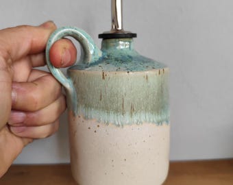 May include: A ceramic oil dispenser with a handle and a stainless steel spout. The stoneware jug features a light blue and green glaze over a cream-colored base. The handle is a matching light blue color.