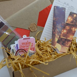 Könnte beinhalten: Ein brauner Karton, gefüllt mit Bastelbedarf, darunter ein Set farbiges Washi-Tape, ein Stickerbogen mit dem Text "main character energy", eine Rolle Washi-Tape und zwei Lesezeichen mit Bildern von Gebäuden. Der Karton ist mit braunem Packpapier gefüllt.