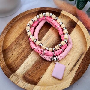 May include: A stack of three bracelets made from flat, circular clay beads in shades of cream, bright pink, and light pink. The bracelets are accented with small, round, metallic silver beads and feature a single rectangular light purple charm.